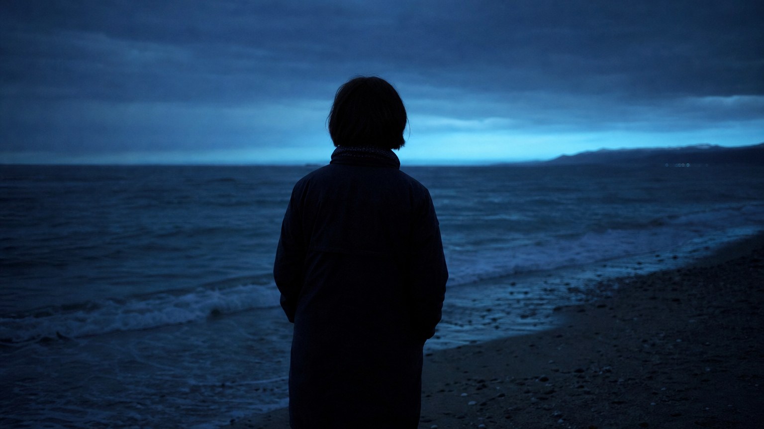 夕暮れの浜辺で海を見つめる人物の後ろ姿(明日への予告ビジュアル)